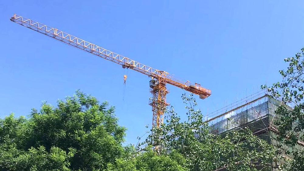 Yellow crane next to a building under construction, surrounded by green trees on a clear day.