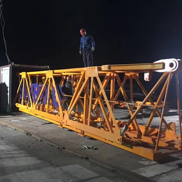 Un ouvrier debout sur une grande poutre industrielle jaune la nuit, à côté d'une grue à tour à montage automatique IHURMO qtz900 tc7082 d'une longueur de flèche de 70 pieds.