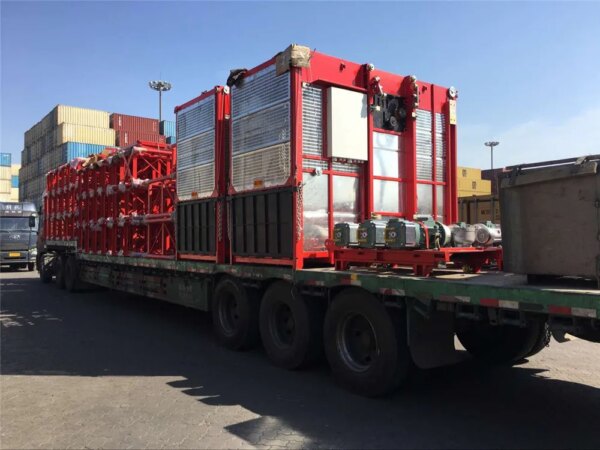 Ein Pritschenanhänger transportiert einen Doppelkäfigaufzug für den Hochbau, im Hintergrund sind Container zu sehen.