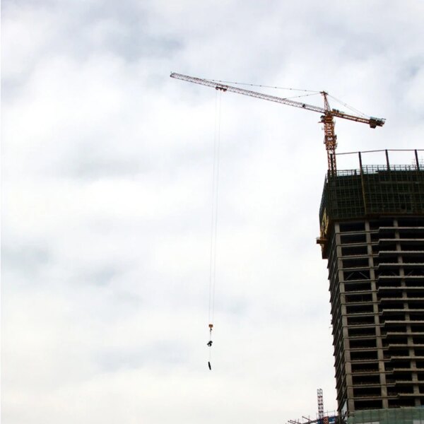 Bouwkraan, met name de IHURMO CE 50 m hoog 10 ton QTZ125 TC6015 torenkraan, opererend naast een hoog gebouw in aanbouw tegen een bewolkte hemel.