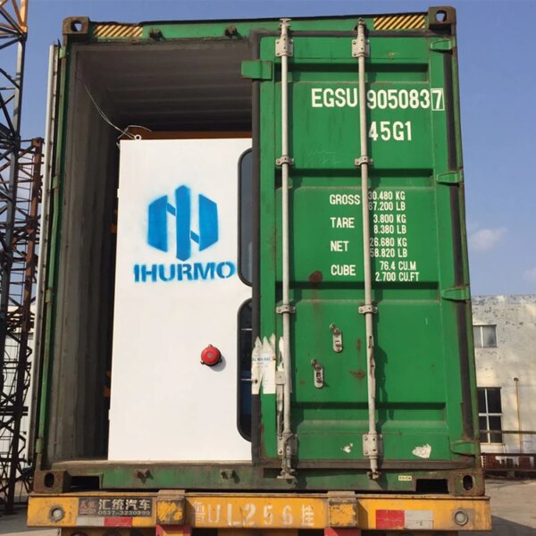 Green shipping container with a blue and white logo, and a red circular object on the door, positioned under an IHURMO CE Standard QTZ50 TC5010 City Tower Crane on a yellow chassis.