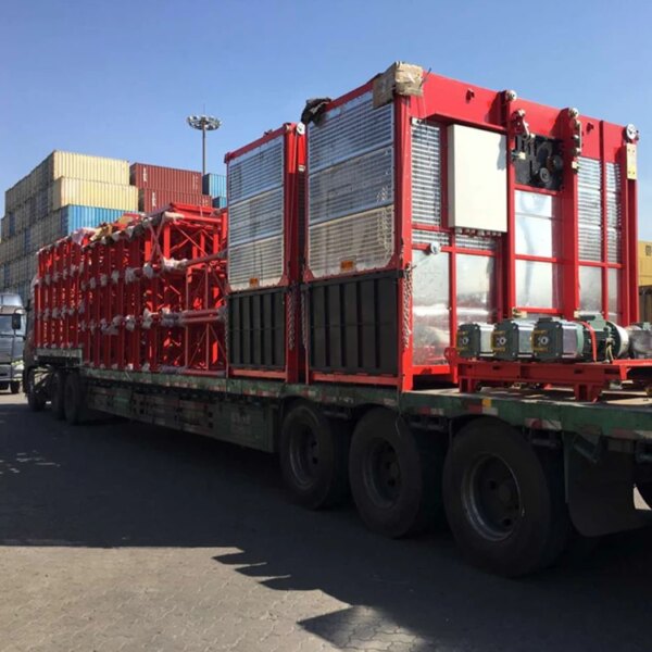 Un camión de plataforma cargado con un elevador de piñón y cremallera de doble jaula para construcción, equipo industrial rojo y contenedores de carga asegurados.