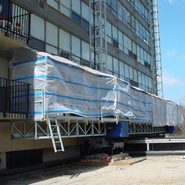 Scaffolding with an IHURMO single-mast mast climber and a protective tarp covering part of a multi-story building under construction or renovation.