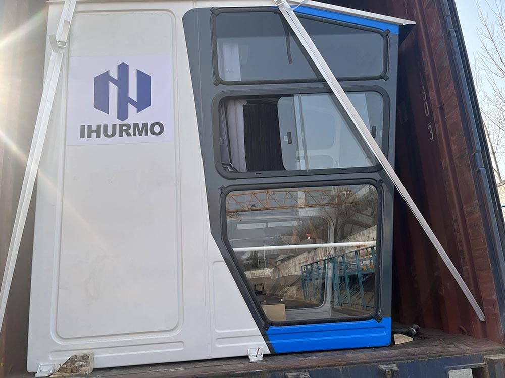 A container holds a large white structure with dark windows, blue base, and "IHURMO" logo, secured by white straps.