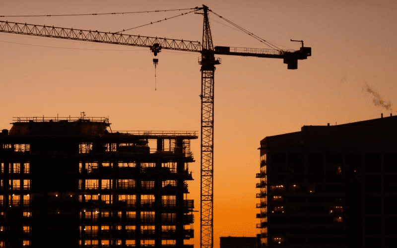 Silhouet van een bouwplaats met een kraan bovenop een wolkenkrabber tegen een oranje zonsondergang.