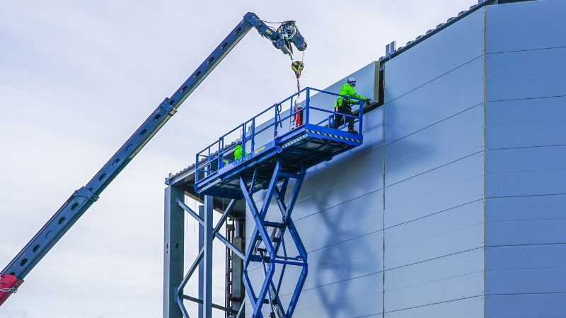 Arbeiter in Sicherheitsausrüstung benutzen eine blaue Scherenbühne für Bauarbeiten an einem großen grauen Industriegebäude. In der Nähe steht ein Kranarm.