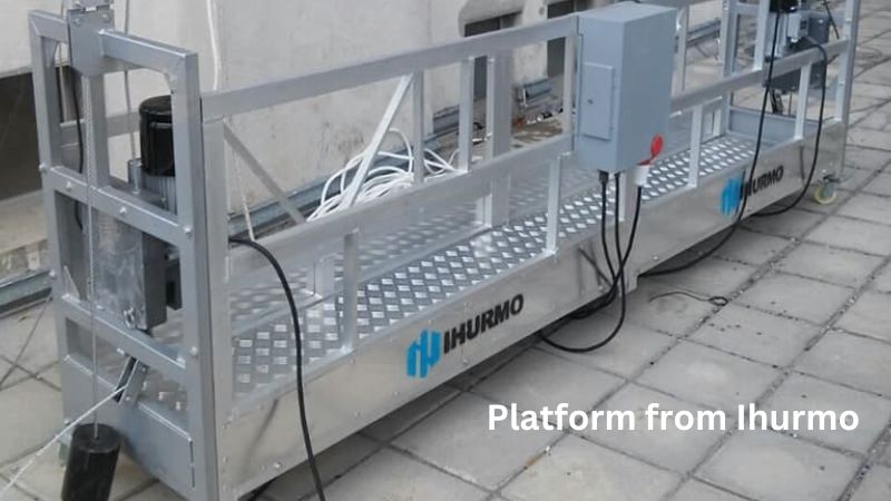 Una plataforma plateada con barandillas y cables sobre una superficie de baldosas presenta el logotipo "IHURMO", lo que garantiza la seguridad para tareas elevadas.