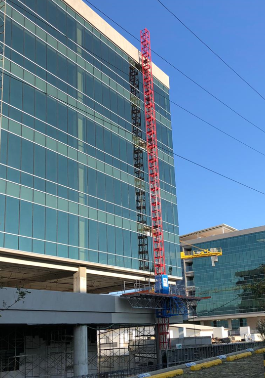 A modern glass office building under construction with a red mast climber. Another IHURMO glass building stands in the clear blue sky.