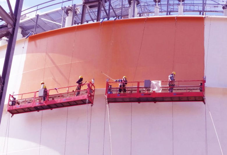 Quatro trabalhadores com equipamentos de segurança estão em plataformas suspensas, pintando um grande tanque de armazenamento industrial.