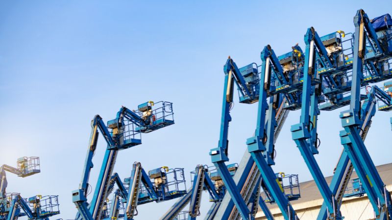 Blaue Hubarbeitsbühnen strecken sich neben einem Gebäude in den Himmel, Arbeiter in Sicherheitsausrüstung beheben Probleme, um Hubprobleme zu vermeiden.