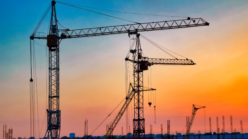 Guindastes em silhueta ao pôr do sol contra um céu azul-alaranjado durante um projeto de construção.