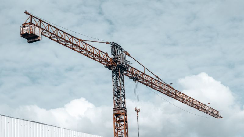 Sebuah menara derek tinggi dengan jib panjang berdiri di depan langit berawan di dekat sebuah bangunan dengan atap bergelombang putih, menggambarkan konsep perencanaan proyek.