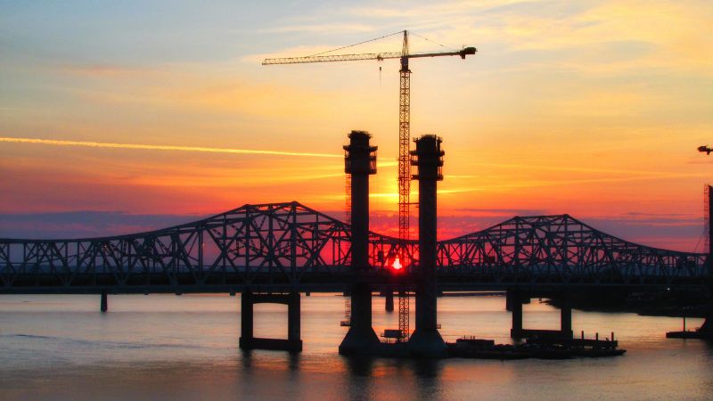 Una scena di costruzione di un ponte al tramonto presenta gru, torri parzialmente costruite e un vecchio ponte sullo sfondo.