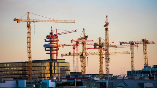 Au coucher du soleil, les grues d'un chantier de construction très fréquenté, dont une grue à tour et une grue mobile, fonctionnent en synchronisation, encadrées par des bâtiments et une tour de communication.