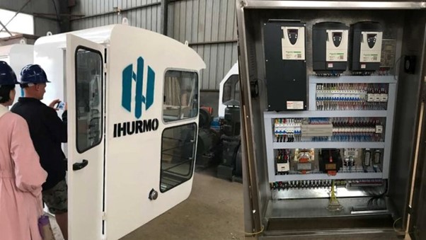 Due persone con caschi protettivi ispezionano una cabina "IHURMO" vicino a un pannello di controllo aperto, fondamentale per il progetto della gru a torre.