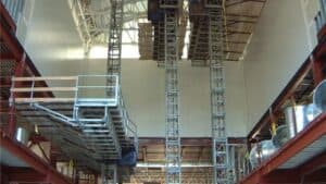 O edifício industrial em construção conta com andaimes e mastros de escalada apoiados em vigas de aço e plataformas metálicas.