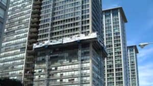 Um arranha-céu com uma plataforma de construção pende em sua fachada, com janelas de vidro e sacadas. Outro edifício é parcialmente visível nas proximidades.