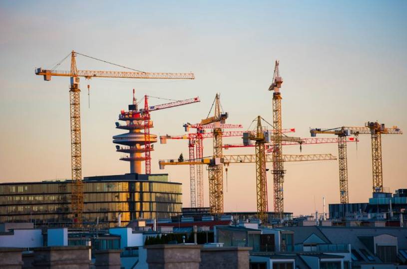 Tower cranes rise above a construction site, with buildings in front and a clear sky behind.
