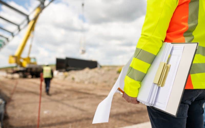 Een arbeider in een veiligheidshesje met documenten houdt een bouwplaats in de gaten met een kraan, een hijskraan en een andere arbeider in de buurt.