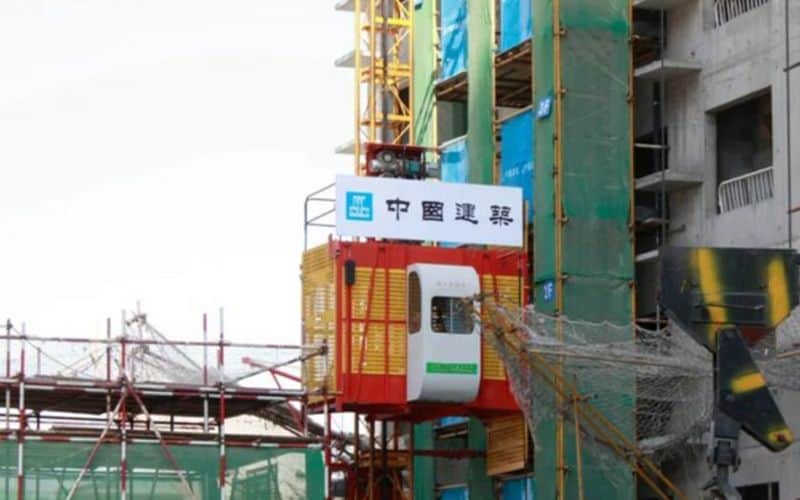 Ascenseur à palan jaune et rouge IHURMO sur un chantier, attaché à un bâtiment avec une signalisation chinoise.
