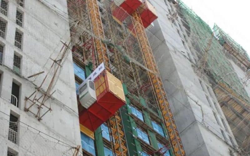 Le monte-charge IHURMO soulève les matériaux et les ouvriers d'un grand bâtiment en construction, avec des échafaudages et des filets de sécurité pour plus de sécurité.