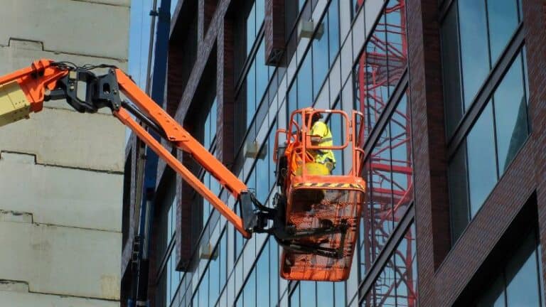 Een arbeider in een veiligheidshesje gebruikt een oranje hoogwerker bij de ramen van een gebouw met meerdere verdiepingen.