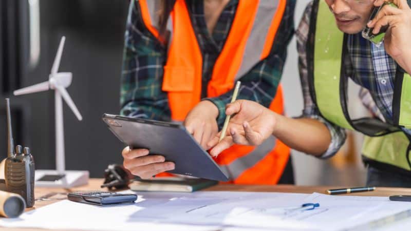 Twee ingenieurs in veiligheidsvesten bekijken plannen op een tablet aan een bureau met blauwdrukken, een windturbinemodel en documenten.
