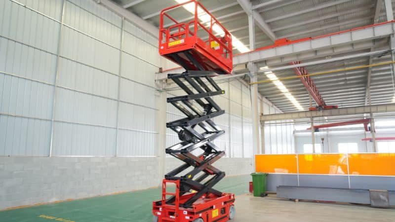 In een ruim magazijn met hoge plafonds en metalen wanden staat een rode hoogwerker volledig uitgeschoven.