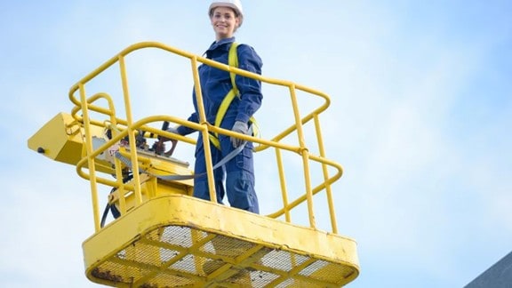 Een persoon in veiligheidsuitrusting en helm staat op een gele hoogwerker onder een helderblauwe lucht.