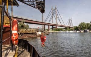 เรือจอดเทียบท่า ตะขอเครนสีแดง และสะพานแขวนขนาดใหญ่ทอดข้ามแม่น้ำ.