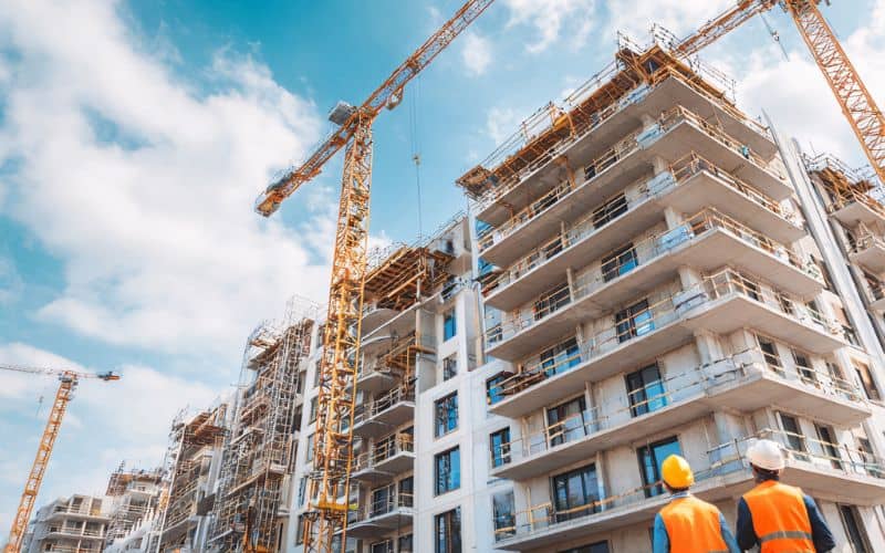 Torenkranen zijn aan het werk boven een gebouw in aanbouw, terwijl twee werknemers in veiligheidskleding de bouwplaats in de gaten houden.