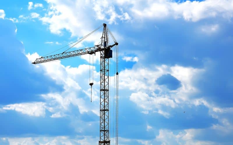 Een bouwkraan steekt hoog af tegen de blauwe, met wolken omhulde lucht, terwijl machinisten behendig kranen op de bouwplaats manoeuvreren.