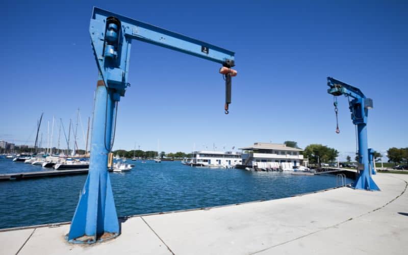 Twee blauwe scheepsliften op een steiger bij een jachthaven, met zeilboten en een gebouw onder een helderblauwe lucht.