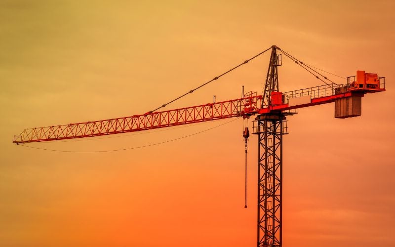 Een kraan met een lange arm steekt af tegen een oranje-gele zonsondergang. Voor het comfort van de machinist boven de grond zijn er toiletten aanwezig.