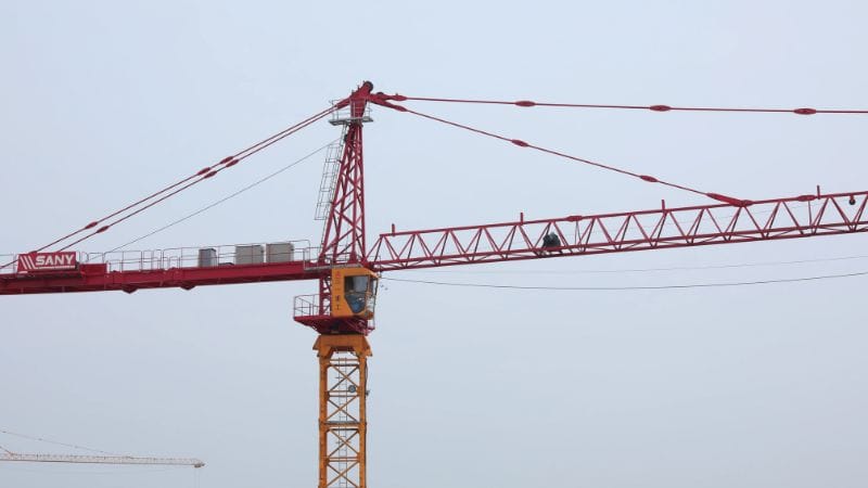 एक व्यक्ति धुंधले, बादलों से घिरे आकाश के बीच एक लाल रंग की निर्माण क्रेन की क्षैतिज भुजा पर चल रहा है।.