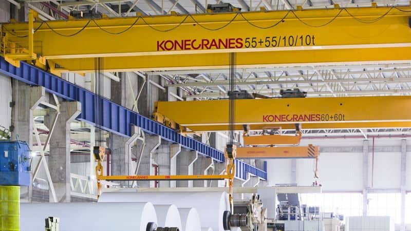 ऊँची छतों वाले एक चमकदार रोशनी से भरे औद्योगिक गोदाम में पीले रंग की कोनेक्रेंस गैन्ट्री क्रेनें बड़े-बड़े सामग्री के रोल उठा रही हैं।.
