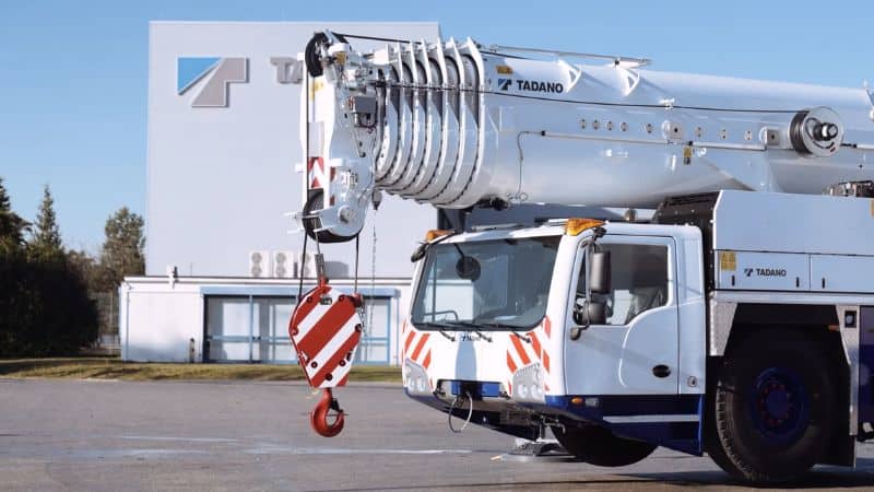 एक शीर्ष 10 निर्माता की लाल-सफेद हुक वाली मोबाइल क्रेन, उपयोग के लिए तैयार अवस्था में, तादानो की एक इमारत के सामने खड़ी है।.