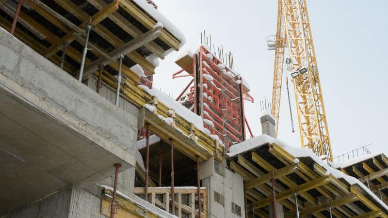 Snow-covered, unfinished concrete building with exposed beams, scaffolding, and a yellow tower crane in the background.