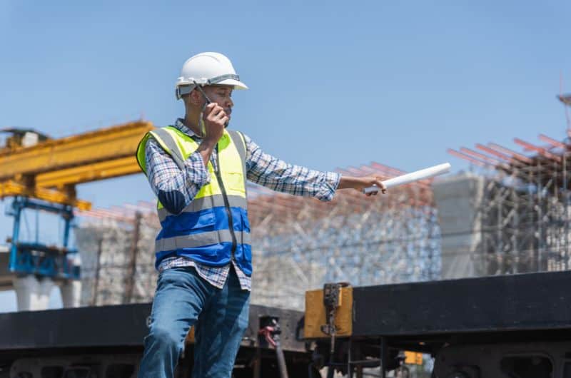 A construction worker in safety gear gestures with a rolled-up paper and holds a walkie-talkie at an outdoor construction site.