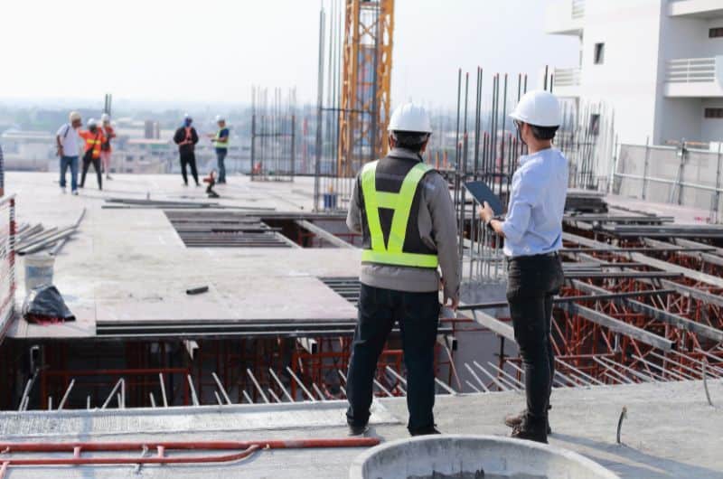 Two construction workers in safety gear stand on a building site, with others and scaffolding visible in the background.