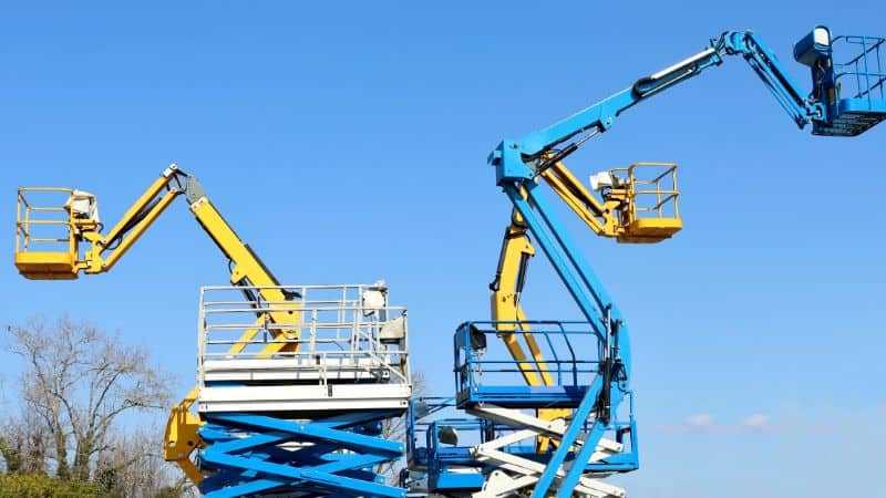 Yellow and blue scissor lifts and cherry pickers are raised against a clear sky, with trees visible in the background.