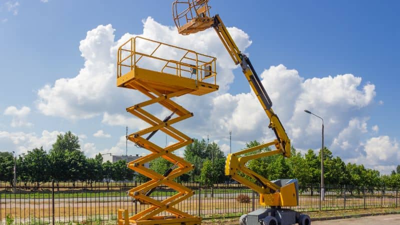 A yellow scissor lift and cherry picker are parked side by side outdoors with trees and a metal fence in the background.