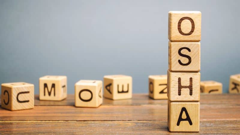 Wooden blocks spell "OSHA" vertically on a table, with scattered blocks and a neutral background.