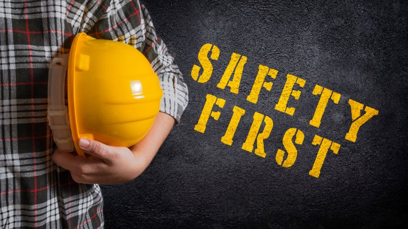 Person holding a yellow hard hat labeled "SAFETY FIRST" on a dark background, emphasizing workplace safety.