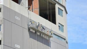 Two construction workers on a suspended scaffold follow OSHA guidelines while working on a multi-story building’s exterior.