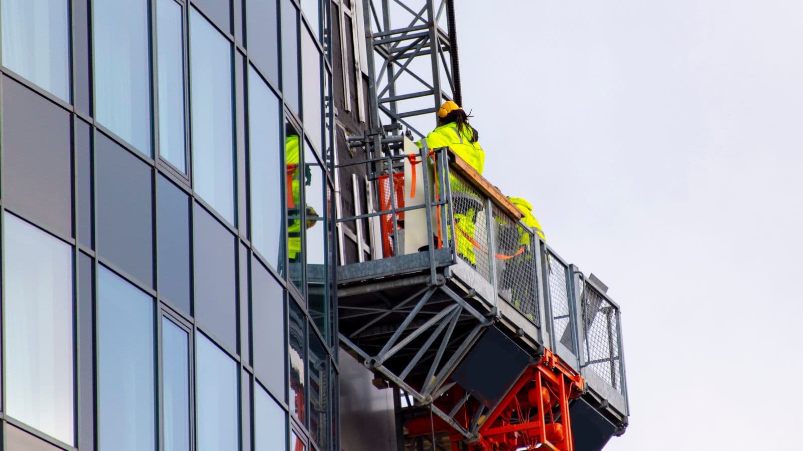 Workers in high-visibility safety gear operating a mast climbing work platform on a modern glass building.