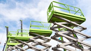 A green scissor lift extended outdoors against a blue sky with clouds, used for construction or maintenance work at height.