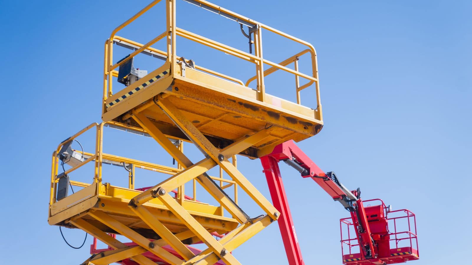 A yellow scissor lift with a red articulated boom against a clear blue sky, demonstrating proper safety features and operation for construction and maintenance work.