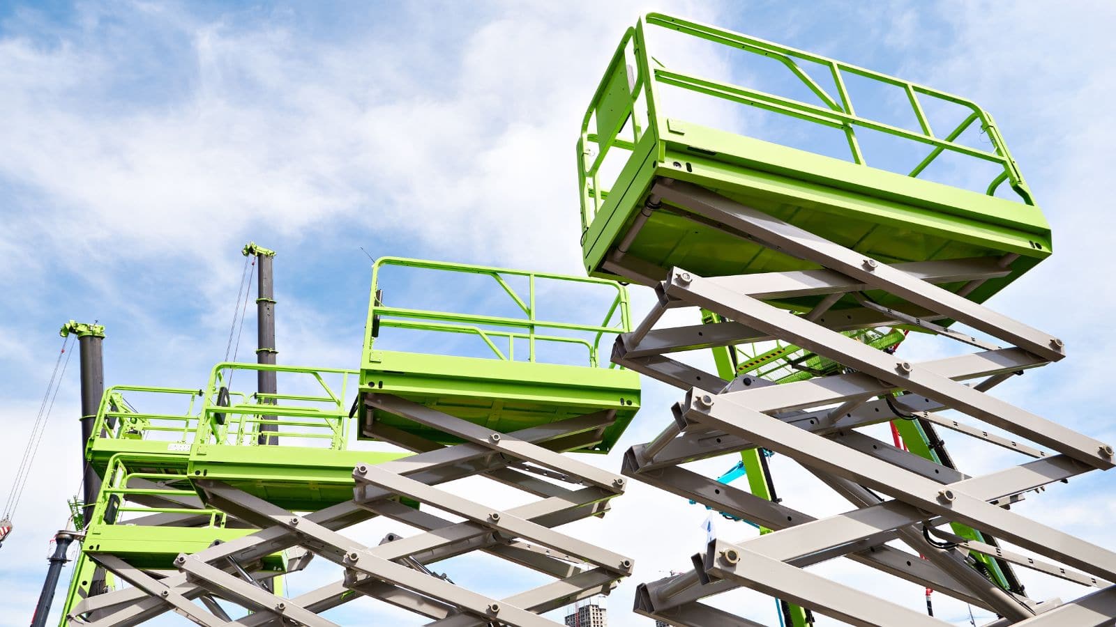 A green scissor lift extended outdoors against a blue sky with clouds, used for construction or maintenance work at height.
