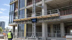 Construction worker in safety gear observing a suspended platform on a building under construction with a tower crane in the background.